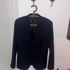 Mörkblå klassisk kavaj - Snygg mörkblå kavaj med två knappar framtill och klassisk krage. Fodrad insida och raka fickor ger en stilren look. Perfekt för dig som vill ha en enkel och tidlös stil. Passar till skjorta eller t-shirt för en clean outfit.