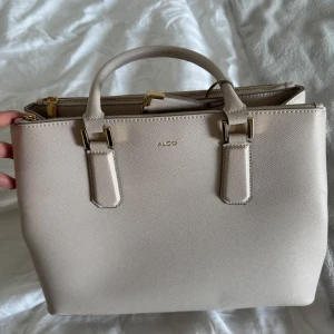 Beige handväska från ALDO med guldiga detaljer - Snygg beige handväska från ALDO med stilrena guldiga metalldetaljer och struktur i materialet. Väskan har både handtag och avtagbar axelrem, flera fack med dragkedja och smarta innerfickor. Köpt för 1200kr, säljer för 450. Pris kan diskuteras. 