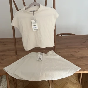 Vit ribbad polotopp från Zara - Säljer en stilren vit polotopp från Zara i ribbad bomull. Toppen har korta ärmar och en högre krage som ger en clean look. Perfekt att matcha med kjol eller jeans för en fräsch outfit. Enkel och snygg design med croppad passform. OBS: Topp i M, Se litet sträck på kjolen på bild 3, vet ej om det går bort i tvätt då settet är oanvänt) 