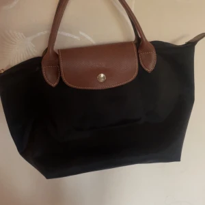 Svart handväska från Longchamp - Säljer pga att den inte kommer till användning längre. Klassisk svart handväska från Longchamp med bruna skinnhandtag och lock. Väskan har guldfärgad dragkedja med Longchamp-logga och är tillverkad i slitstark syntet med detaljer i skinn. Perfekt storlek för vardagens essentials och enkel att matcha. Den är i den lilla storleken, inga defekter, super fin 