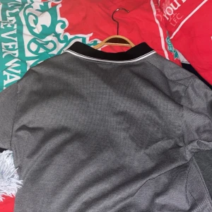 Svartvit pikétröja från Hugo Boss - Snygg svartvit pikétröja från Hugo Boss med diskret rutigt mönster. Tröjan har svart krage med vita ränder, knäppning framtill och korta ärmar. Perfekt för dig som gillar stilrena och klassiska plagg med modern passform.