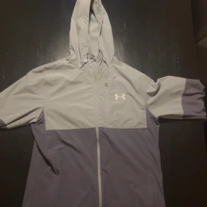 Grå vindjacka från Under Armour - Snygg tvåfärgad vindjacka från Under Armour i ljusgrått och mörkgrått. Jackan har huva med dragsko, hel dragkedja framtill och diskret logga på bröstet. Perfekt för blåsiga dagar och sportiga outfits.