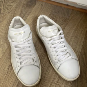 Vita Saint Laurent sneakers i skinn - Klassiska vita sneakers från Saint Laurent Paris i äkta skinn. Snygg minimalistisk design med rund tå, platt sula och diskret guldtext på plösen. Perfekta för dig som gillar stilrena och lyxiga skor med premiumkänsla.