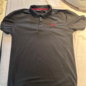 Svart pikétröja från Under Armour - Svart pikétröja från Under Armour med röd logga på bröstet och i nacken. Klassisk krage och knäppning med två knappar. Tröjan är kortärmad och har en clean, sportig look som funkar till många stilar.
