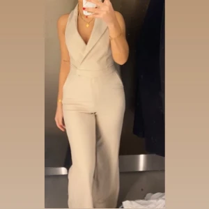 Ny och oanvänd Beige omlott jumpsuit med v-ringning - Snygg beige jumpsuit med omlott och djup v-ringning. Vida ben och ärmlös design ger en stilren och modern look. Perfekt för dig som vill sticka ut med en elegant och trendig outfit.