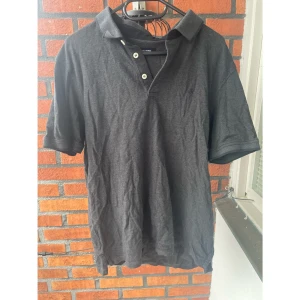 Mörkgrå pikétröja från Jack & Jones storlek L  - Snygg mörkgrå pikétröja från Jack & Jones med klassisk krage och tre vita knappar framtill. Tröjan har korta ärmar och är tillverkad i mjuk bomull storlek saknas men skulle säga att den passar L 