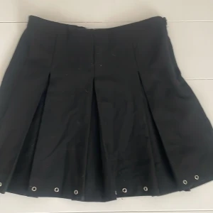 Svart plisserad kjol från Zara - Svart kortkjol från Zara med plisserad design och coola metallöljetter längst ner. Kjolen har justerbar midja med knapp på insidan och dragkedja i sidan. Perfekt för dig som gillar en edgy och stilren look.