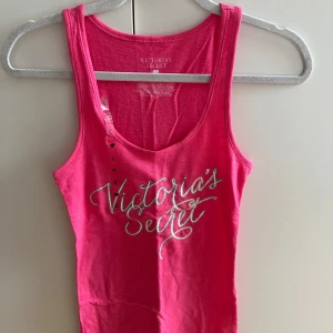 Rosa linnetopp från Victoria's Secret - Snygg rosa linnetopp från Victoria's Secret med glittrigt silvrigt tryck framtill. Modellen är ärmlös med rund halsringning och har en figurnära passform. Materialet är en mjuk mix av bomull, polyester och elastan som känns skön huden