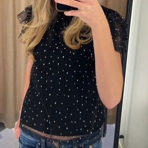 Svart blus med stenar och volangärm - Supersöt svart blus med transparent mesh och små stenar över hela. Blusen har volangärmar i flera lager. Perfekt för dig som gillar detaljer och vill sticka ut lite extra.