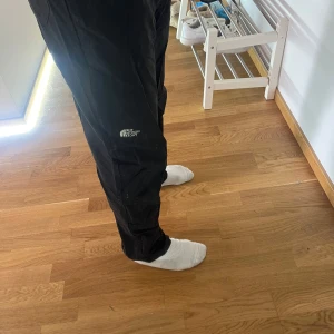 Svarta täckbyxor från The North Face - Svarta täckbyxor från The North Face med logga på benet. Byxorna har en loose passform och är perfekta för kalla dagar. Tillverkade i slitstarkt syntetmaterial och har en sportig look med elastiska benslut.