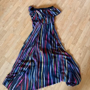 Randig långklänning från Fervente - Säljer en färgglad långklänning från Fervente med offshoulder och volang upptill. Klänningen har ett snyggt randigt mönster i blått, rött, gult, grönt och lila. Materialet känns lätt och flowy, perfekt för varma dagar. Resår upptill för skön passform.