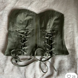 Olivgrön korsett med snörning - Snygg olivgrön korsett med snörning framtill och dragkedja i mitten. Axelbandslös modell med markerad midja och coola detaljer. Perfekt om du ska på fest eller bara vill vara lite uppklädd i vardagen. Oanvänd och jättesnygg. Skriv om du har frågor. Pris kan diskuteras. Den är i storlek M, men jag skulle säga att den även passar S.