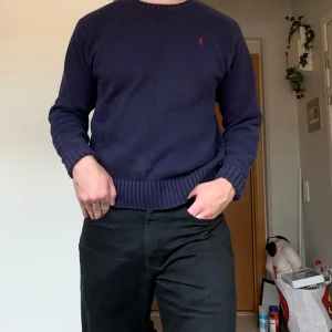 Vintage Stickad Ralph Lauren - Klassisk mörkblå stickad tröja från Polo Ralph Lauren i 100% bomull. Tröjan har rund hals, ribbade muddar och nederkant samt den ikoniska röda loggan broderad på bröstet. Heavyknit och riktigt slitstark. Tyvärr har den en blekt fläck som säkert går o fixa på framsidan men ingenting jag själv brukar lägga märke till. Sitter som en M