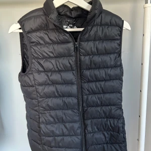 Svart dunväst med dragkedja - Snygg svart dunväst med dragkedja framtill. Perfekt för lager-på-lager under kyliga dagar. Västen har en stilren design med quiltade detaljer och hög krage för extra värme.