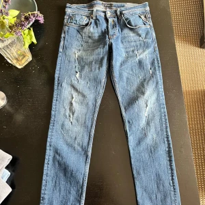 Replay blå slitna jeans, stl 32/32 - Säljer ett par blå jeans från Replay. Jeansen har snygga slitningar framtill, klassisk femficksdesign och raka ben. Materialet är jeans med orangea kontrastsömmar och detaljerade bakfickor. Perfekta för en avslappnad och trendig look.