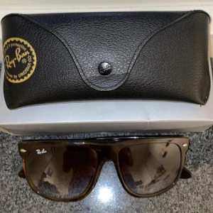 Ray-Ban bruna Wayfarer solglasögon - Snygga Ray-Ban Wayfarer solglasögon med brunmelerad båge och mörka tonade glas. Klassisk fyrkantig form och diskret Ray-Ban-logga på båda sidor. Kommer med original svart fodral i läderlook. Perfekta för soliga dagar och en cool stil.