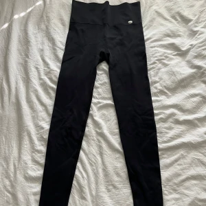 Svarta höga ribbade träningstights - Svarta ribbade leggings med hög midja och diskret metallogo framtill. De är tighta och stretchiga, perfekta för träning eller chill.