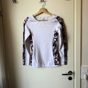 Vit träningströja Stronger XS - Supersnygg vit långärmad träningströja från Stronger i storlek XS. Tröjan har blommiga och lila detaljer längs sidorna och ärmarna, samt meshpartier upptill på ryggen. Perfekt för dig som vill sticka ut på gymmet eller löprundan! Storlek XS. Fint skick, men mer använd. 