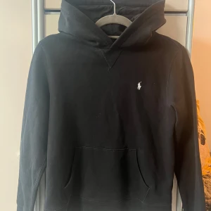 Svart hoodie från Polo Ralph Lauren - Svart hoodie från Polo Ralph Lauren med klassisk huva och känguruficka. Liten vit broderad logga på bröstet. Mjuk bomullskänsla och långärmad modell, perfekt för chill dagar eller när du vill ha en clean look.