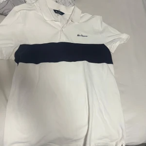 Vit pikétröja Ben Sherman med blå rand - Snygg vit pikétröja från Ben Sherman med en bred marinblå rand över bröstet och klassisk krage. Tröjan har korta ärmar och broderad logga på bröstet. Perfekt för en clean och sportig stil.