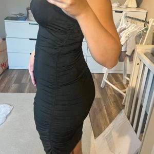 Svart tight omlottklänning - Säljer en svart, tight klänning med omlott-effekt och smala axelband. Klänningen är kort och har snygga rynkade detaljer längs sidorna som framhäver figuren. Perfekt för dig som gillar en stilren och trendig look.