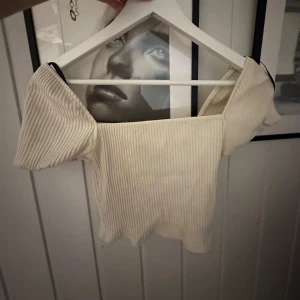Ribbad offshoulder topp i beige - Superfin croppad offshoulder topp i ribbad beige bomull. Toppen har korta ärmar och en vågig kant nertill som ger en extra snygg detalj. Perfekt att styla med höga jeans eller kjol för en trendig look.