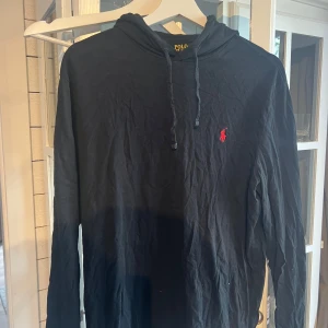 Svart långärmad tröja från Polo Ralph Lauren - Svart tröja från Polo Ralph Lauren i mjuk bomull. Klassisk design med huva och snörning, samt det ikoniska röda broderade logotypen på bröstet. Långärmad och perfekt för en chill och stilren look. Är inte tjock som en hoodie utan som en långärmad T-shirt med huva. 