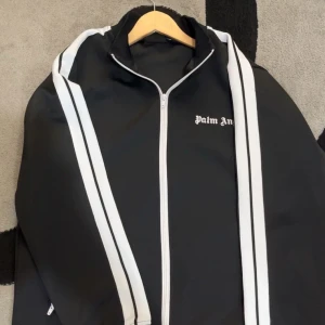 Palm Angels svart track jacket L - Svart track jacket från Palm Angels med vita ränder längs ärmarna och vit dragkedja. Jackan har logga på bröstet och sportig vibe. Tillverkad i polyester och bomull med elastan för stretch. Perfekt för dig som gillar streetwear och snygga detaljer.
