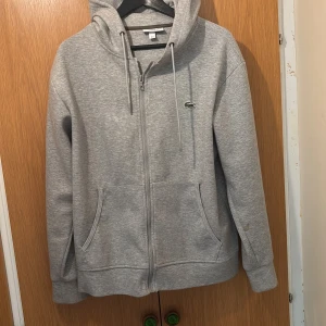 Grå hoodie från Lacoste med dragkedja - Snygg grå hoodie från Lacoste, säljer för jag använder aldrig den. Den är i perfekt skick, fläckfri och knappt använd.
