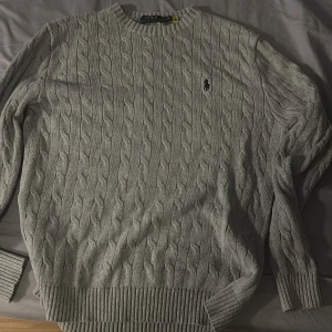 Grå kabelstickad tröja Polo Ralph Lauren - Kabelstickad grå tröja från Polo Ralph Lauren med klassisk rund halsringning och svart broderad logga på bröstet. Tröjan har ribbade muddar och är tillverkad i mjuk bomull. Perfekt för lager-på-lager och snygg till jeans.