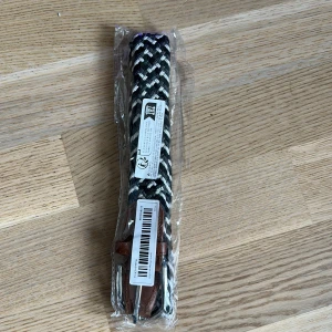 Svart och vit flätat bälte - Snyggt flätat bälte i svart och vitt med bruna skinn-detaljer vid spännet. Bältet har ett randigt mönster och är perfekt för att ge outfiten en cool touch. Passar till både jeans och chinos.