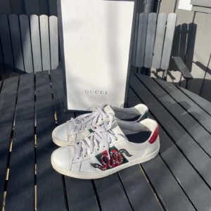 Gucci Ace snake - Gucci Ace sneakers i vitt skinn med ikonisk broderad röd och svart orm på sidan. Baksidan har metallicröd och grön detalj. Klassisk låg modell med snörning och rund tå. Perfekta för dig som vill sticka ut med lyxig streetstyle.