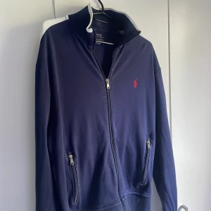 Mörkblå zip från Polo Ralph Lauren - Mörkblå zip från Polo Ralph Lauren med dragkedja framtill och två fickor med dragkedja. Klassisk röd logga broderad på bröstet. Perfekt för dig som gillar stilren och sportig look.