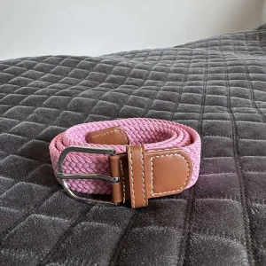 Flätat rosa bälte med skinn-detaljer - Snyggt flätat bälte i ljusrosa med bruna skinn-detaljer och silvrigt spänne. Perfekt accessoar för att lyfta din outfit med en pop av färg. Bältet har en klassisk design med kontrastsömmar på skinnet och är både stilrent och trendigt.