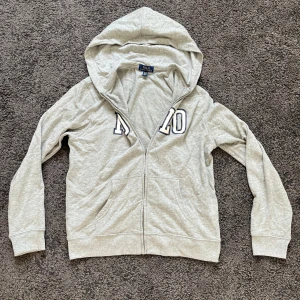 Grå hoodie från Polo Ralph Lauren - Snygg grå hoodie från Polo Ralph Lauren med dragkedja och huva. Stor POLO-logga i vitt och svart på bröstet. Mjuk bomullskänsla, två fickor fram och långa ärmar. Perfekt för chill dagar eller när du vill ha en clean streetstyle.