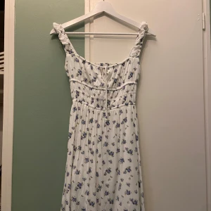 Vit blommig kortklänning med knyt - Supersöt vit kortklänning med blått blommönster och breda axelband. Klänningen har smock i midjan, knytning framtill och lätt rynkad kjol. Från stradivarius. 
