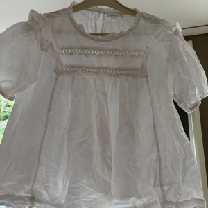 Vit romantisk blus med spetsdetaljer - Supersöt vit blus från Gina Tricot med puffiga korta ärmar, volangdetaljer vid axlarna och spetsband framtill. Blusen har rund halsringning och knäppning bak i nacken. Perfekt för dig som gillar en romantisk och luftig stil.