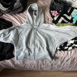 Grå Adidas hoodie med dragkedja - Snygg grå hoodie från Adidas med klassiska vita ränder på ärmarna och dragkedja framtill. Tröjan har huva med snörning och två fickor. Perfekt för en sportig och chill stil. Adidas-logga på bröstet och detaljer i silver på dragkedjan. Den är croppad både i ärmar o mage