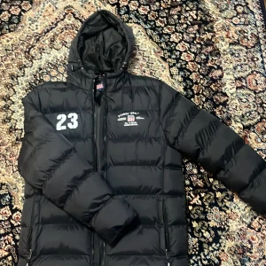 Svart pufferjacka från Vinson, M - Svart pufferjacka från Vinson med huva, dragkedja och vita tryckdetaljer som '23' och logga på bröstet. Jackan är vadderad och har sportig vibe, perfekt för kalla dagar. Snygga detaljer och klassisk amerikansk stil.