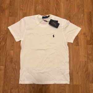 Vit t-shirt från Polo Ralph Lauren - Vit klassisk t-shirt från Polo Ralph Lauren med broderad logga på bröstet. Enkel och stilren design med rund hals och korta ärmar. Tillverkad i mjuk bomull som känns skön mot huden. Perfekt för en clean och tidlös look.