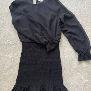 Svart kortklänning från KappAhl - Svart kortklänning från KappAhl med långärmade, transparenta ärmar och smockad midja samt kjol. Klänningen har volangdetalj nertill och en enkel rund halsringning med liten öppning bak. Perfekt för dig som gillar stilren och trendig look.