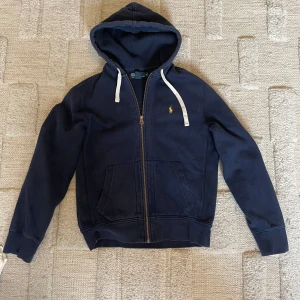 Mörkblå hoodie från Polo Ralph Lauren - Mörkblå hoodie från Polo Ralph Lauren med dragkedja, huva och vita snören. Klassisk gul broderad logga på bröstet och två fickor framtill. Tillverkad i mjuk bomull, perfekt för chill dagar eller när du vill ha en clean streetstyle.