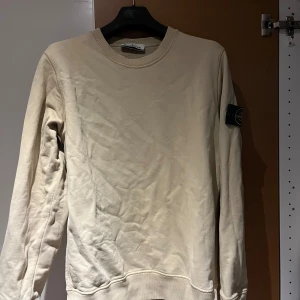 Beige Stone Island sweatshirt - Snygg beige sweatshirt från Stone Island med klassisk rund hals och ikonisk patch på vänster ärm. Tröjan är i mjuk bomull och har ribbade muddar vid ärmslut och nederkant. Perfekt för dig som gillar streetwear och vill ha en clean look.