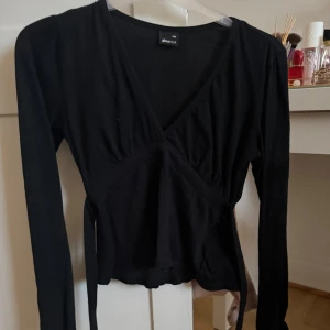 Svart omlottopp från Gina Tricot - Säljer en svart topp med knytning baktill från Gina Tricot i storlek XXS. Toppen har långa ärmar, v-ringning och knytband för en snygg passform. 