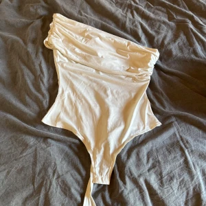Vit asymmetrisk baddräkt - Snygg vit boddy med asymmetrisk axel och draperad överdel. Modellen har en axelbandslös design med en smal axelrem på ena sidan. Materialet är stretchigt och sitter tight mot kroppen, perfekt för fest. Om man vill ha bilder på skriv privat. Aldrig använd 