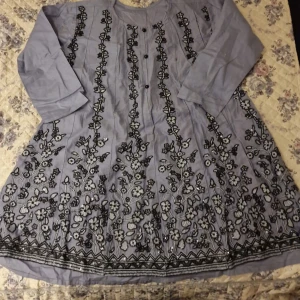 Ljusgrå blommig klänning med broderi - Säljer en ljusgrå klänning med lång ärm och vackert svart blommigt broderi framtill. Klänningen har rund halsringning och ett luftigt fall, perfekt för dig som gillar unika detaljer och mönster. Ej använd. 