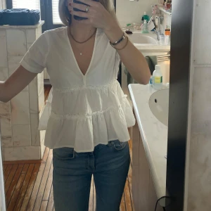 Vit blus med volanger och v-ringning - Säljer en söt vit blus med korta ärmar och v-ringning. Blusen har volangdetaljer och en luftig passform som ger en romantisk känsla. Perfekt att matcha med jeans eller kjol.