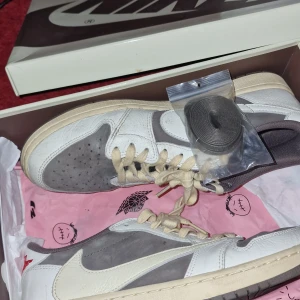 Nike Air Jordan 1 Low Travis Scott x Fragment - Nike Air Jordan 1 Low Travis Scott x Fragment i vitt, grått och beige med ikonisk omvänd swoosh. Skorna har platt sula, rund tå och levereras med extra skosnören. Perfekt för dig som gillar unika sneakers och streetwear.