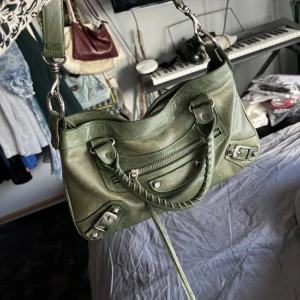 Grön Balenciaga handväska i skinn - Säljer en ikonisk grön handväska från Balenciaga i mjukt skinn. Väskan har både handtag och axelrem, coola silverdetaljer, dragkedja framtill och snygga spännen. Perfekt storlek för det viktigaste och ger en edgy vibe till din outfit. Den mindre modellen, kontakta mig för mått och mer detaljer 💯