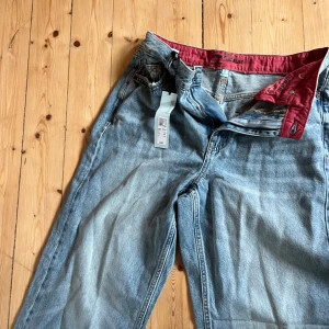 Ljusblåa wide jeans med röd detalj - Säljer ett par ljusblåa jeans med bred passform och snyggt röd foder på insidan av linningen. Jeansen har klassisk femficksdesign, dragkedja och knapp framtill. Perfekta för en avslappnad och trendig look.